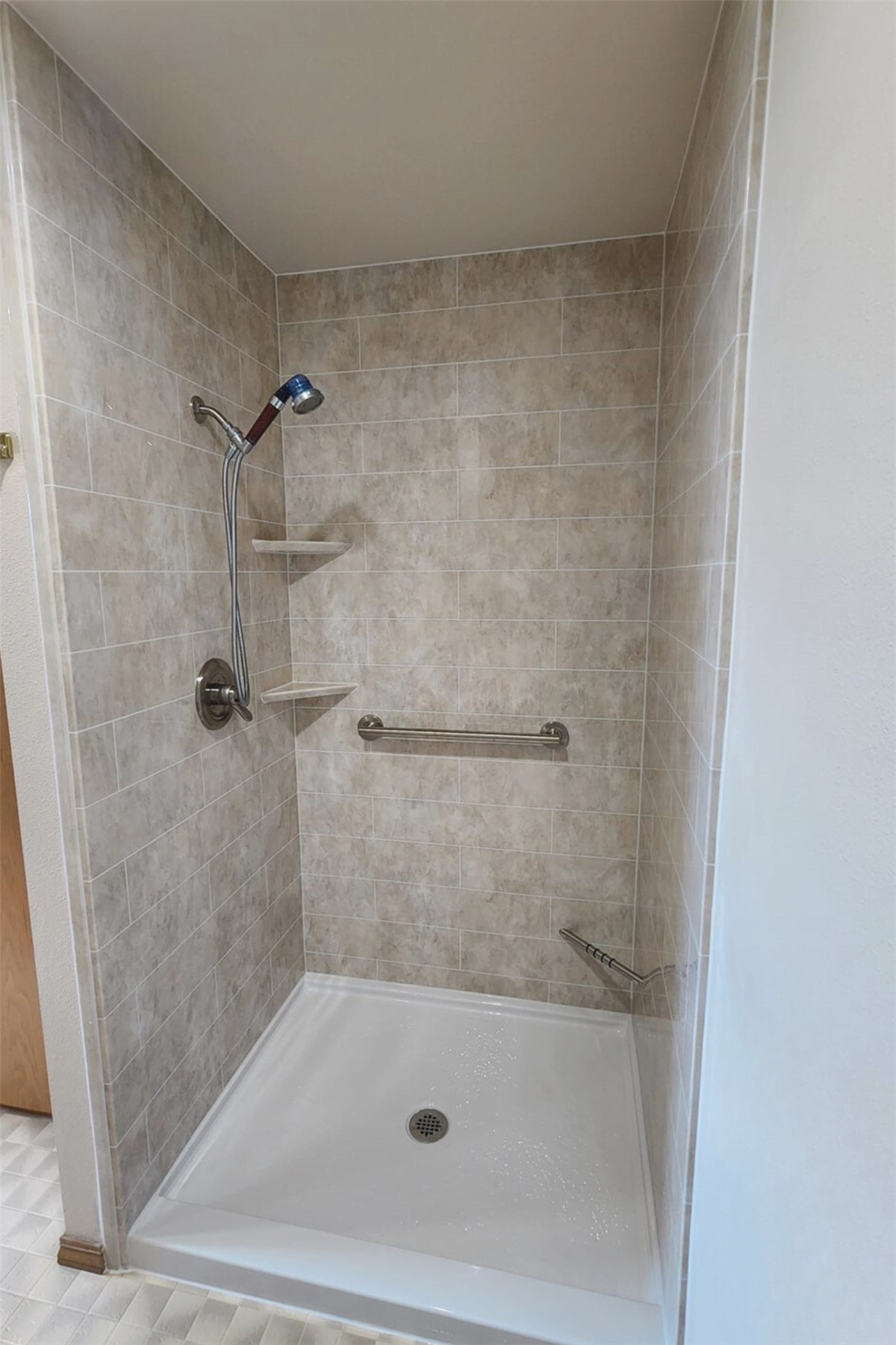 Beige tile-look shower with corner shelves, handheld showerhead, and horizontal grab bar